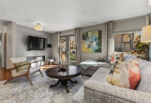 Ein modernes Wohnzimmer mit Kamin, einem wandmontierten Flachbildfernseher, einem runden Couchtisch und einem Sofa mit gemusterten Kissen.