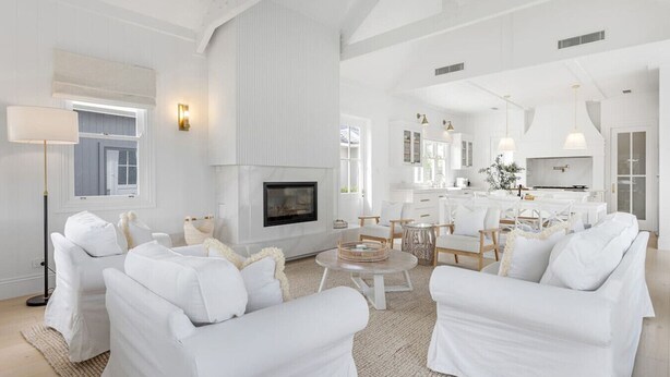 Ein geräumiges Wohnzimmer mit weißen Möbeln, einem Kamin und einer Küchenzeile im Hintergrund.
