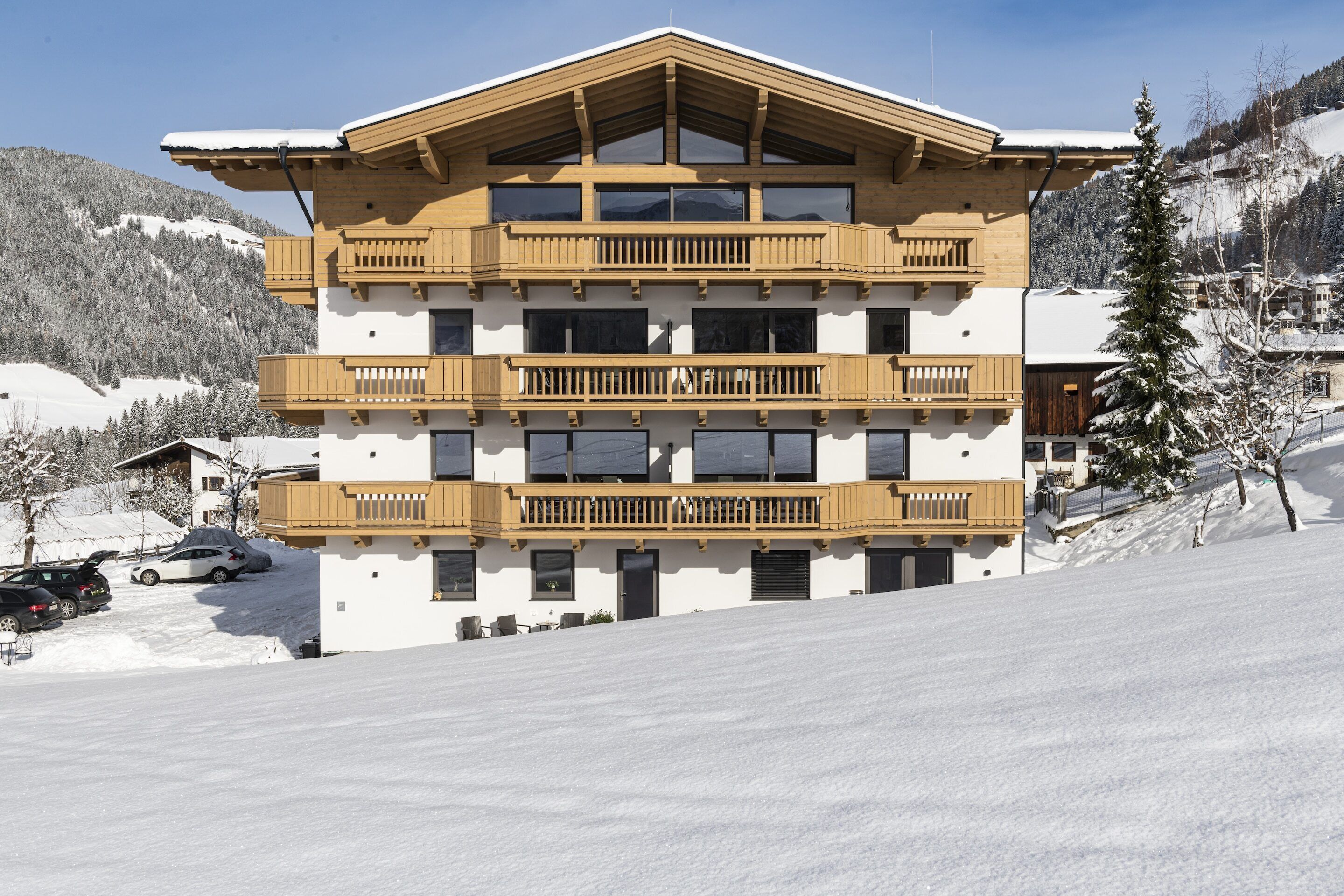 Un edificio de varios pisos con balcones de madera, una pista de esquí al fondo y cielo despejado.