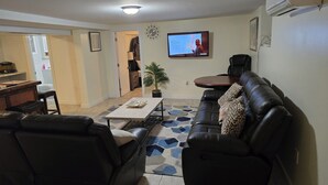 Ein Wohnzimmer mit einer Couch, einem Couchtisch und einem an der Wand montierten Fernseher. Im Hintergrund ist eine Küchenzeile erkennbar.
