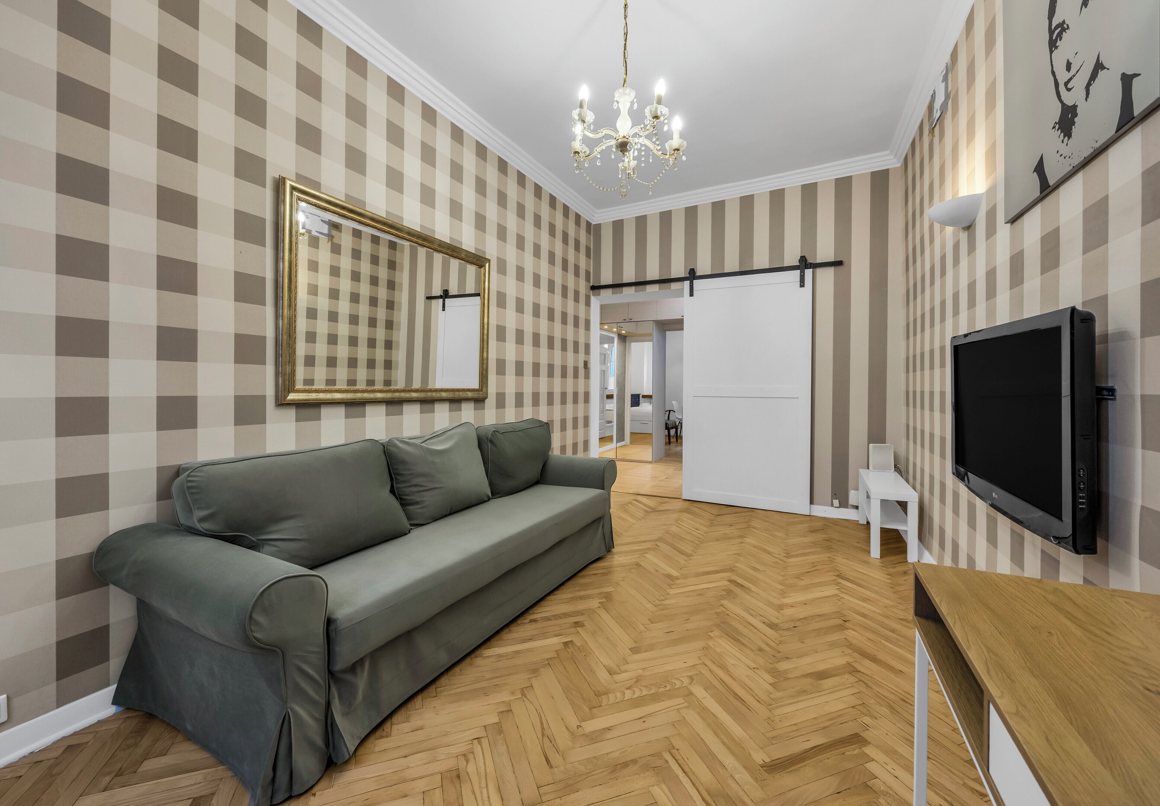 Una sala de estar moderna con suelo de madera en patrón chevron, un sofá gris, un espejo grande, una araña y un televisor de pantalla plana.