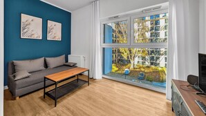 En moderne stue med en grå sofa, et træbord og et stort vindue med udsigt til en boligbygning.