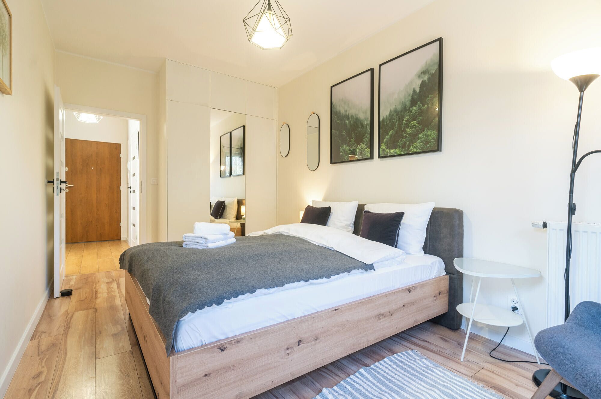 Une chambre à coucher moderne avec un lit en bois, une petite table ronde et une chaise. On y trouve une armoire avec un miroir et une porte qui donne sur une autre pièce.