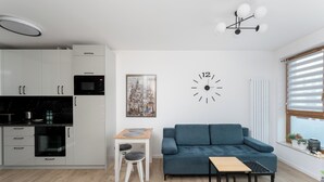 Une cuisine moderne avec des meubles blancs, un four à micro-ondes et une table en bois avec des tabourets. On y trouve également un canapé bleu et une petite table avec un cadre noir. Une grande horloge murale et un tableau encadré sont également visibles.