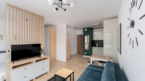 Un salon moderne avec un téléviseur à écran plat, un canapé bleu, un meuble TV en bois, une petite table basse et un coin cuisine à l’arrière-plan.