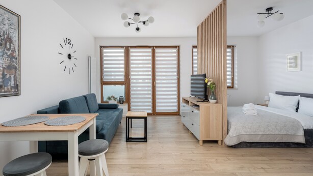 Une pièce moderne et bien éclairée, avec un plancher en bois, un canapé bleu, une table à manger en bois entourée de tabourets, un lit recouvert d’une couette blanche et un téléviseur posé sur un meuble en bois.