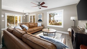 A spacious living room with a brown leather sofa, a ceiling fan, a round coffee table, and a view of the outdoors through large windows. - The Long Lake Escape (Traverse City)