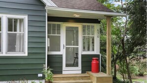 Una casa verde con puerta y ventanas blancas, un porche de madera y un barril rojo.