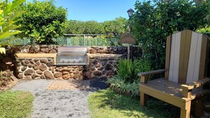 A stone barbecue grill with a stone wall backdrop, a wooden Adirondack chair, and a paved patio area.
