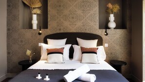 A neatly made bed with white and brown pillows, a dark blue bedspread, and a rolled white towel on the bed. There are two bedside tables with small vases and a wall-mounted light fixture. - Caracalla Room with private patio (Bordeaux)