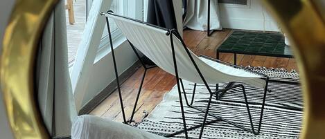 A room with a hammock, a small table with a plant, and a view of the outdoors through a window with curtains.