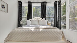 Een bed met een geborduurde witte hoes, zwart gordijn met een geometrisch patroon, en een raam met uitzicht op bomen.