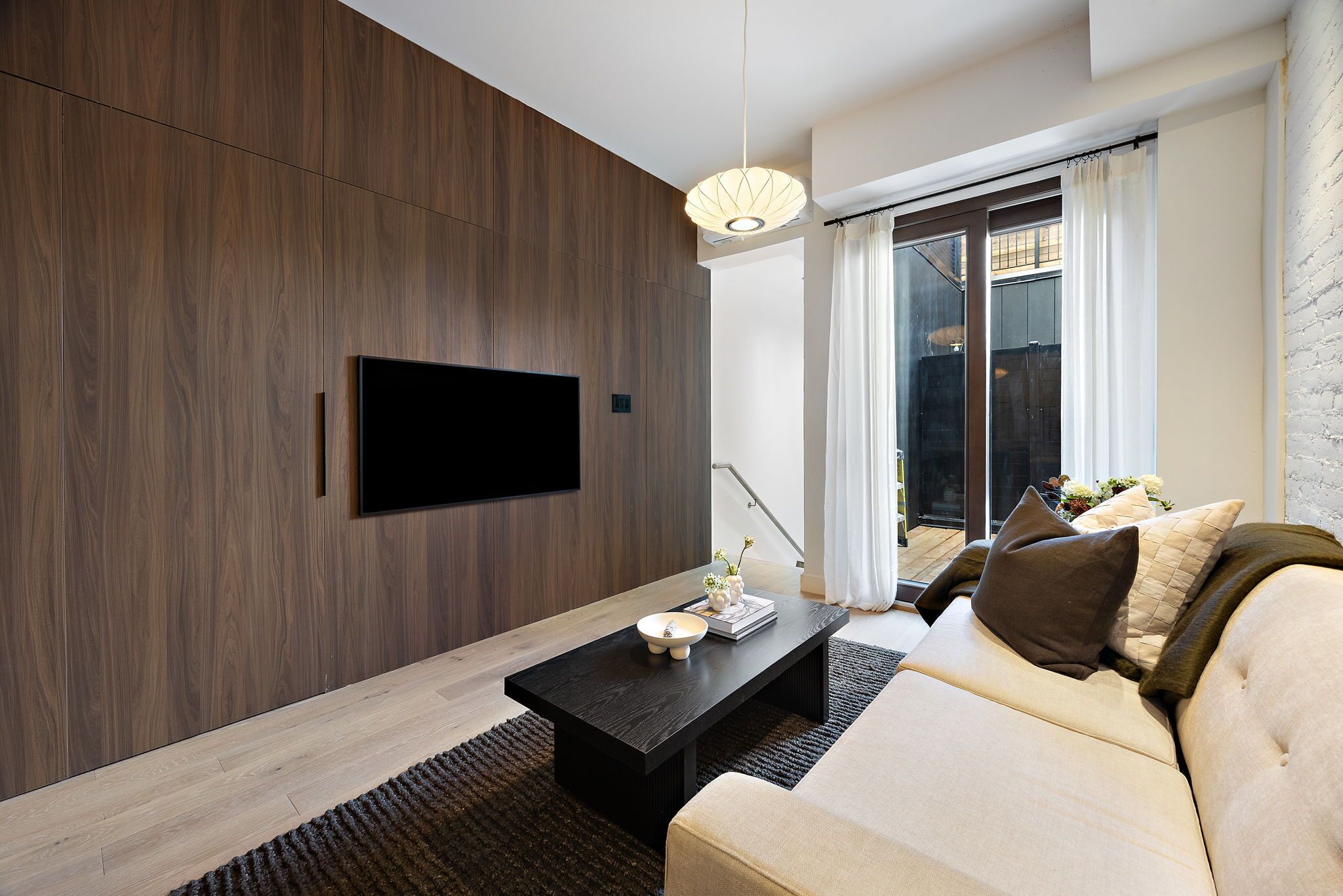A modern living room with a flat-screen TV mounted on a wooden wall, a dark coffee table with decorative items, a beige sofa with cushions, and a large window with curtains.