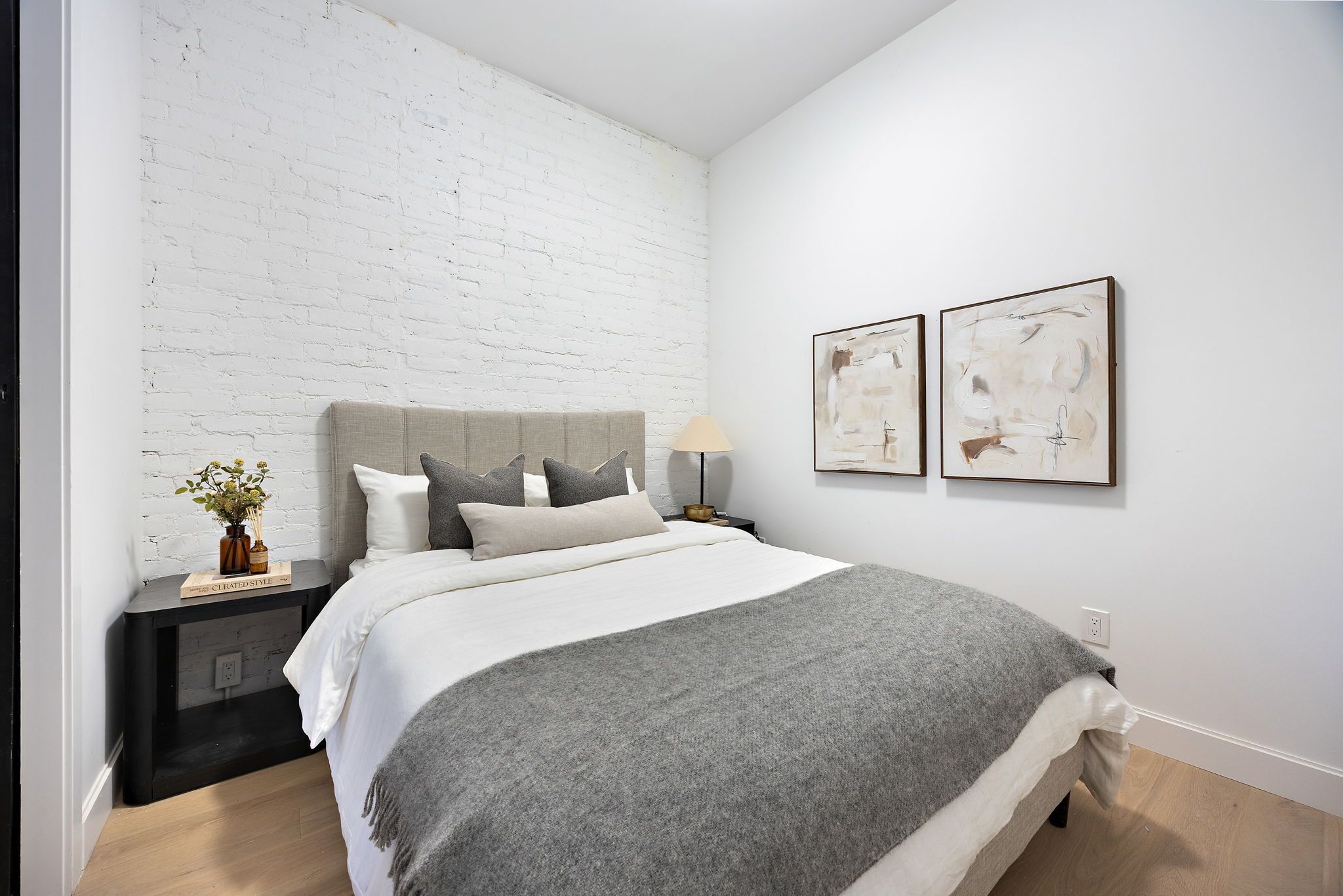 Un dormitorio moderno con una pared de ladrillo blanco, una colcha gris y dos cuadros abstractos en la pared.