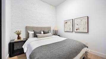 Un dormitorio moderno con una pared de ladrillo blanco, una colcha gris y dos cuadros abstractos en la pared.