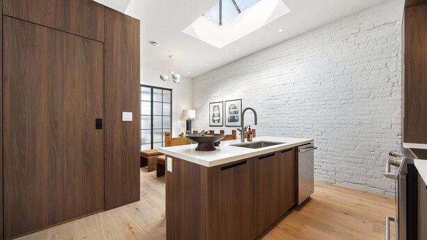 A modern kitchen with a central island, wooden cabinetry, a white brick wall, and a skylight. - The Brick Hotel (Toronto)