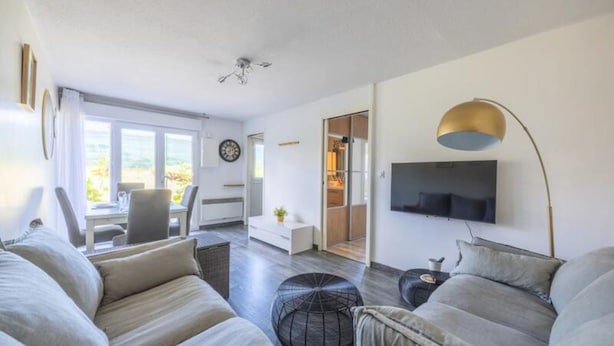 A modern living room with a sofa, ottoman, and a TV mounted on the wall. - Apartment in Villers-sur-mer Near Beach (Villers-sur-Mer)