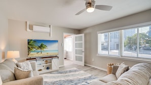 A living room with a large flat-screen TV, a ceiling fan, and a view of the outdoors through large windows. - Across from the beach - Bella by the Sea by SeaBreeze Vacation (Bradenton Beach)