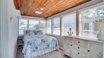 A bedroom with a wooden ceiling, a bed with a blue floral bedspread, a white dresser, and large windows with blinds.