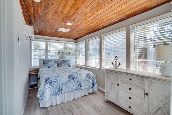 Un dormitorio con techo de madera, una cama con colcha floral azul, un tocador blanco y ventanas grandes con persianas.