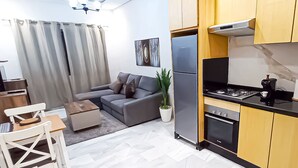 Una sala de estar moderna con un sofá gris, una silla blanca, una mesita y una zona de cocina con estufa, horno y refrigerador.