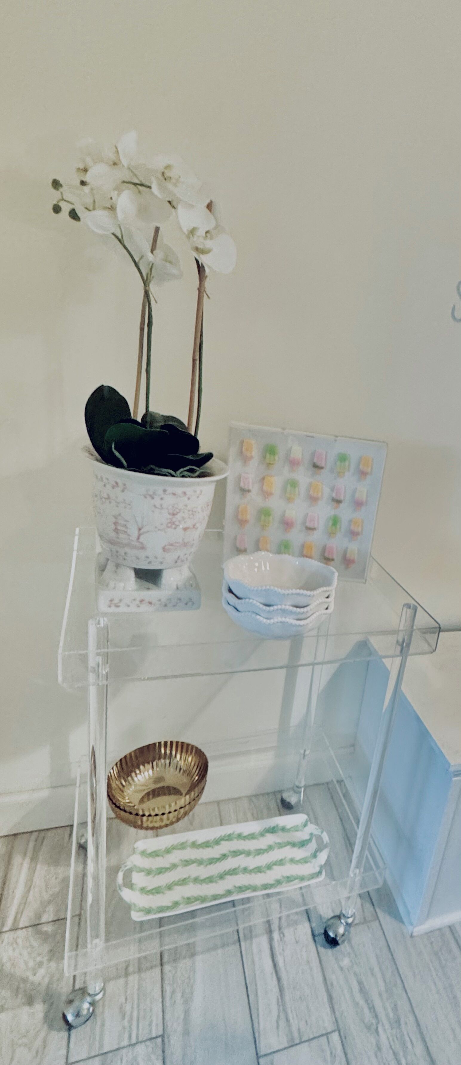 Une étagère en verre transparent sur laquelle sont posés un pot d’orchidée, un bol décoratif et un tableau encadré.