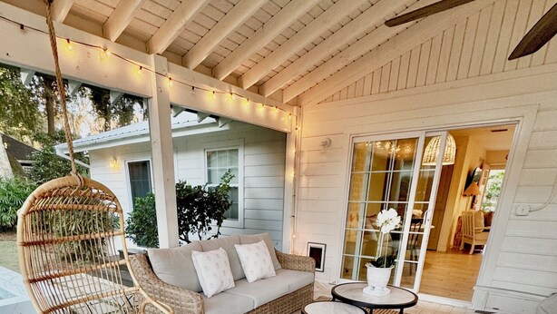 A covered outdoor seating area with a wicker sofa, two round tables, and a hanging chair. - Private Cottage Oasis w Pretty Porch + Splash Pool (St Simons Island)