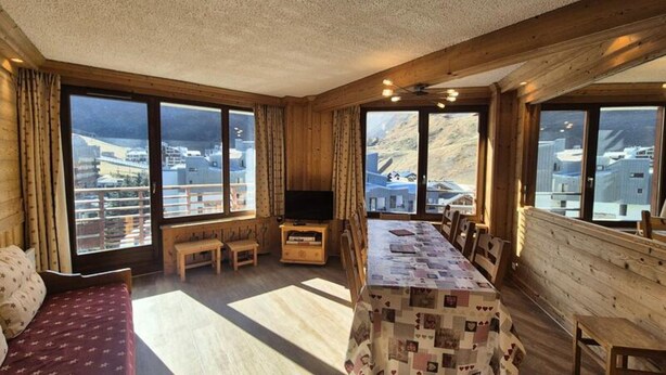 A cozy room with a wooden interior, a large window with a view of a snowy landscape, a dining table with a quilt, and a television on a wooden stand. - Apartment in the Val Claret Area (Tignes)