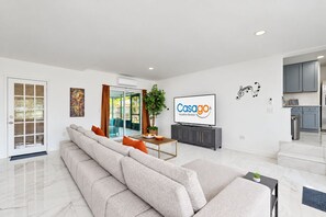 A modern living room with a sofa, a dining table with chairs, a TV, and a kitchen area with cabinets and a countertop. - Stylish Avondale Home – Pet-Friendly & Walkable (Jacksonville)
