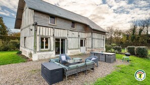 Een huis met twee verdiepingen, een schuin dak, een terras met tuinmeubels en een oprit van grind.
