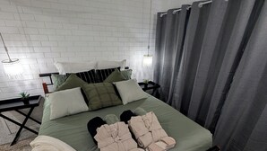 Siististi pedattu sänky, jossa on vihreä päiväpeite, useita tyynyjä ja pari paria tossuja.