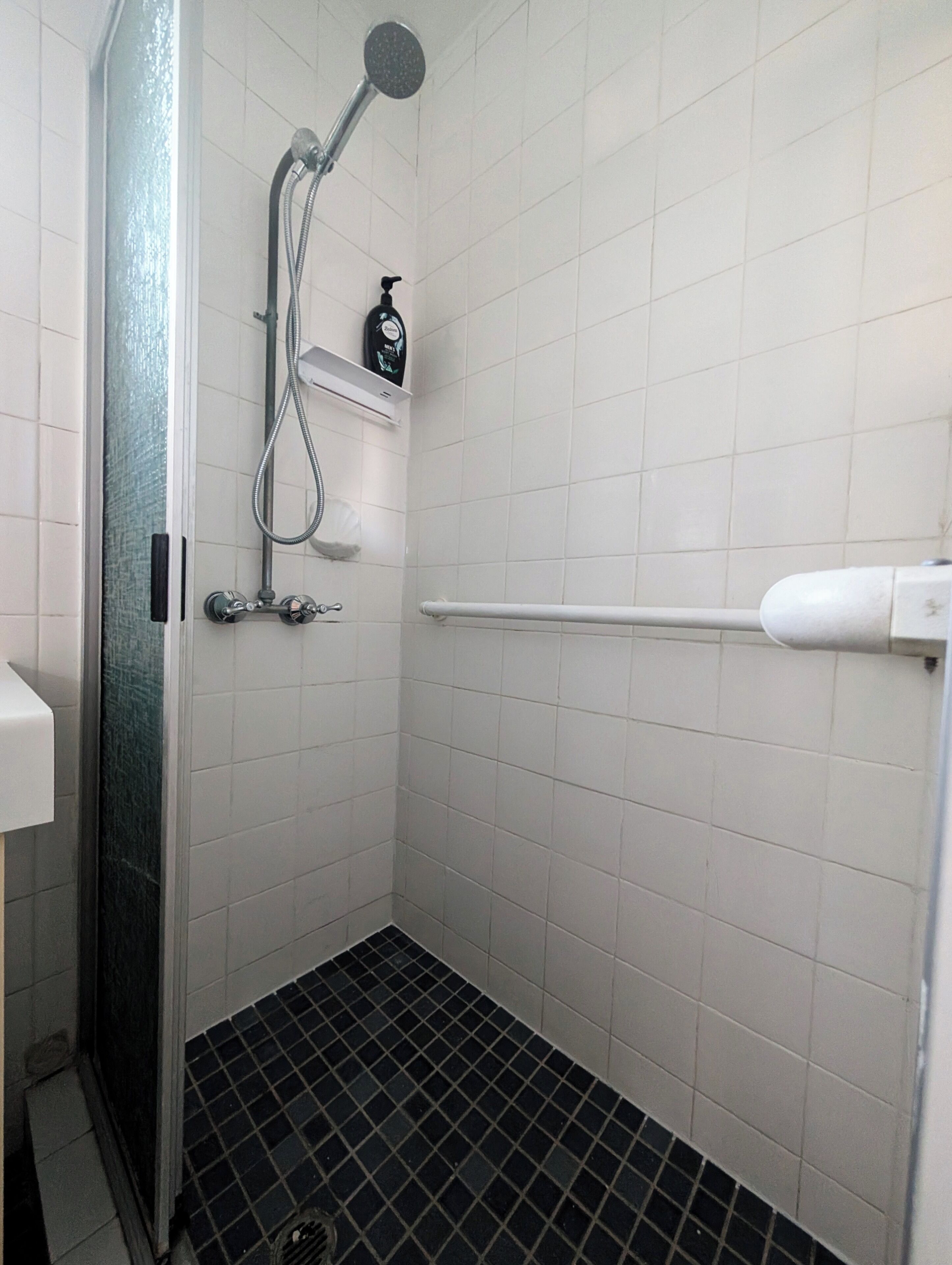 Un baño pequeño con azulejos, un cabezal de ducha manual, un accesorio de ducha manual y un dispensador de jabón en un estante.
