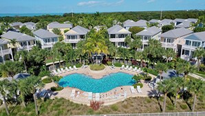 Un resort con una piscina central rodeada de sillones de descanso y palmeras, con varios edificios al fondo.
