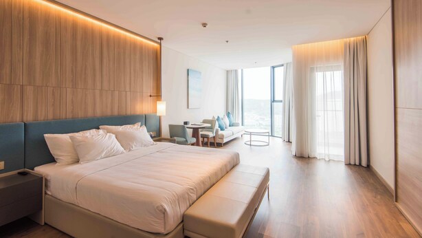 Ein modernes Hotelzimmer mit einem großen Bett, einer Sitzecke mit Tisch und Stühlen und Blick auf die Stadt durch große Fenster.