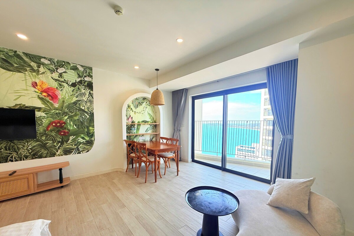 A modern living room with a dining area, a large window with a view, and a tropical-themed wall.