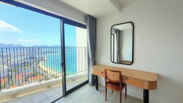 Une pièce comprenant un bureau en bois, une chaise, un miroir et une grande fenêtre donnant sur une ville côtière et des montagnes.