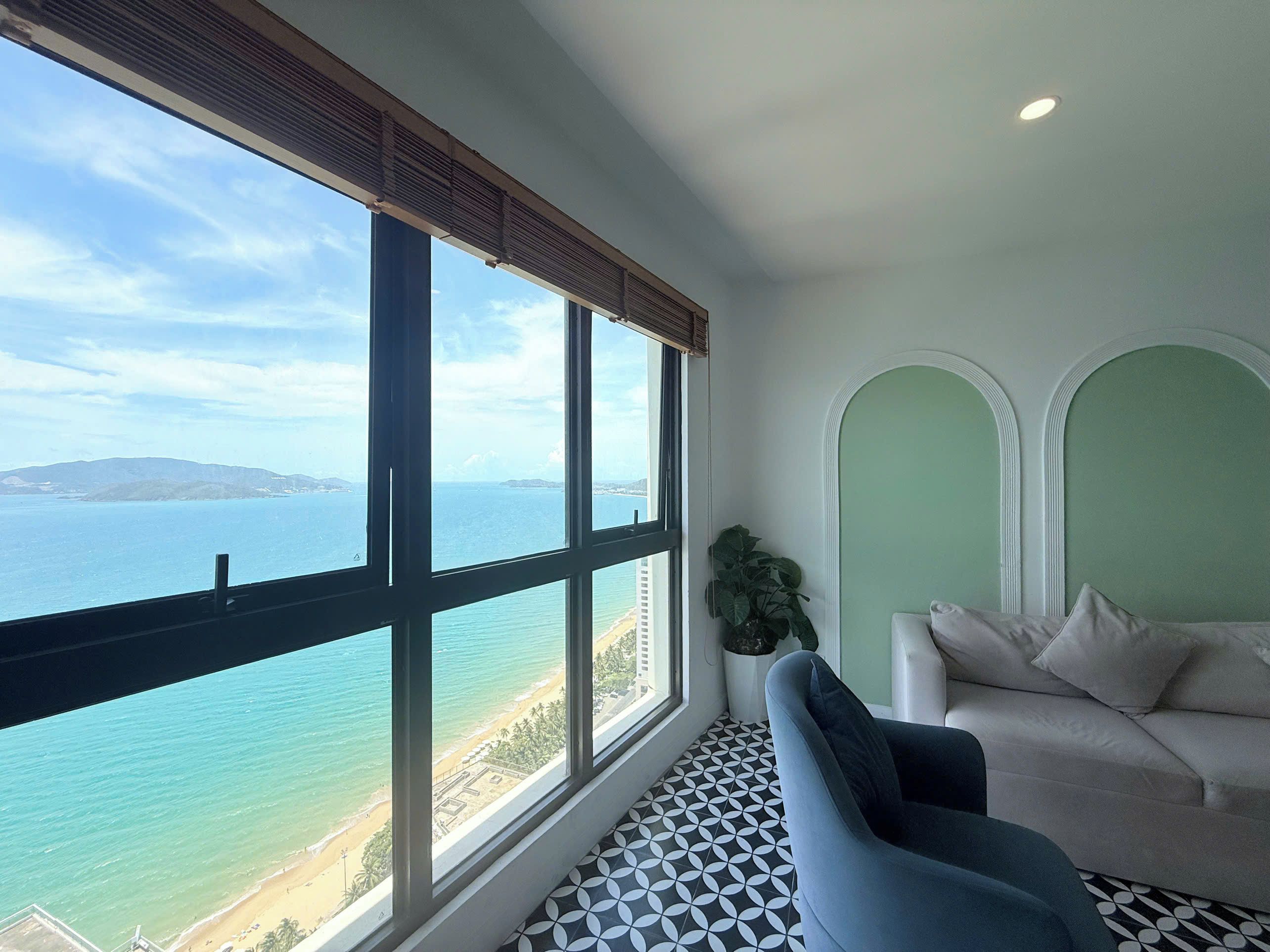 Ein Zimmer mit großem Fenster, das auf einen Strand und das Meer blickt, einer Couch und einer Topfpflanze.
