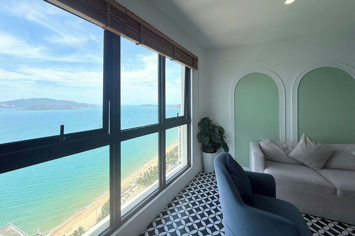 A room with a large window overlooking a beach and the sea, a sofa, and a potted plant.