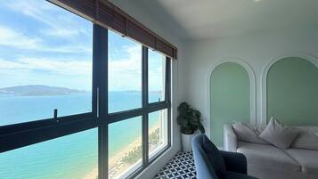 Ein Zimmer mit großem Fenster, das auf einen Strand und das Meer blickt, einer Couch und einer Topfpflanze.