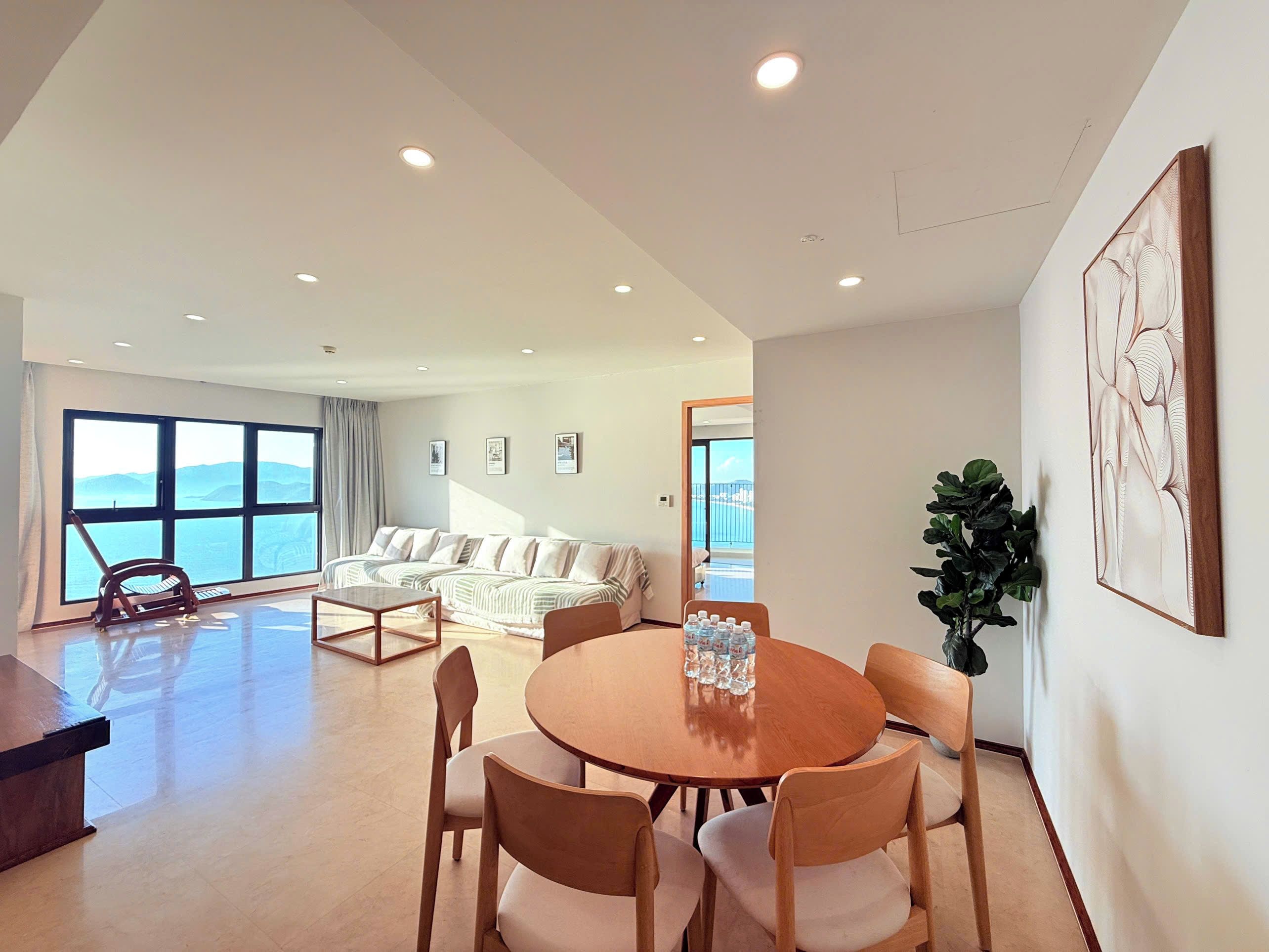 A spacious living area with a large window, a sofa set, a dining table with chairs, and a potted plant.