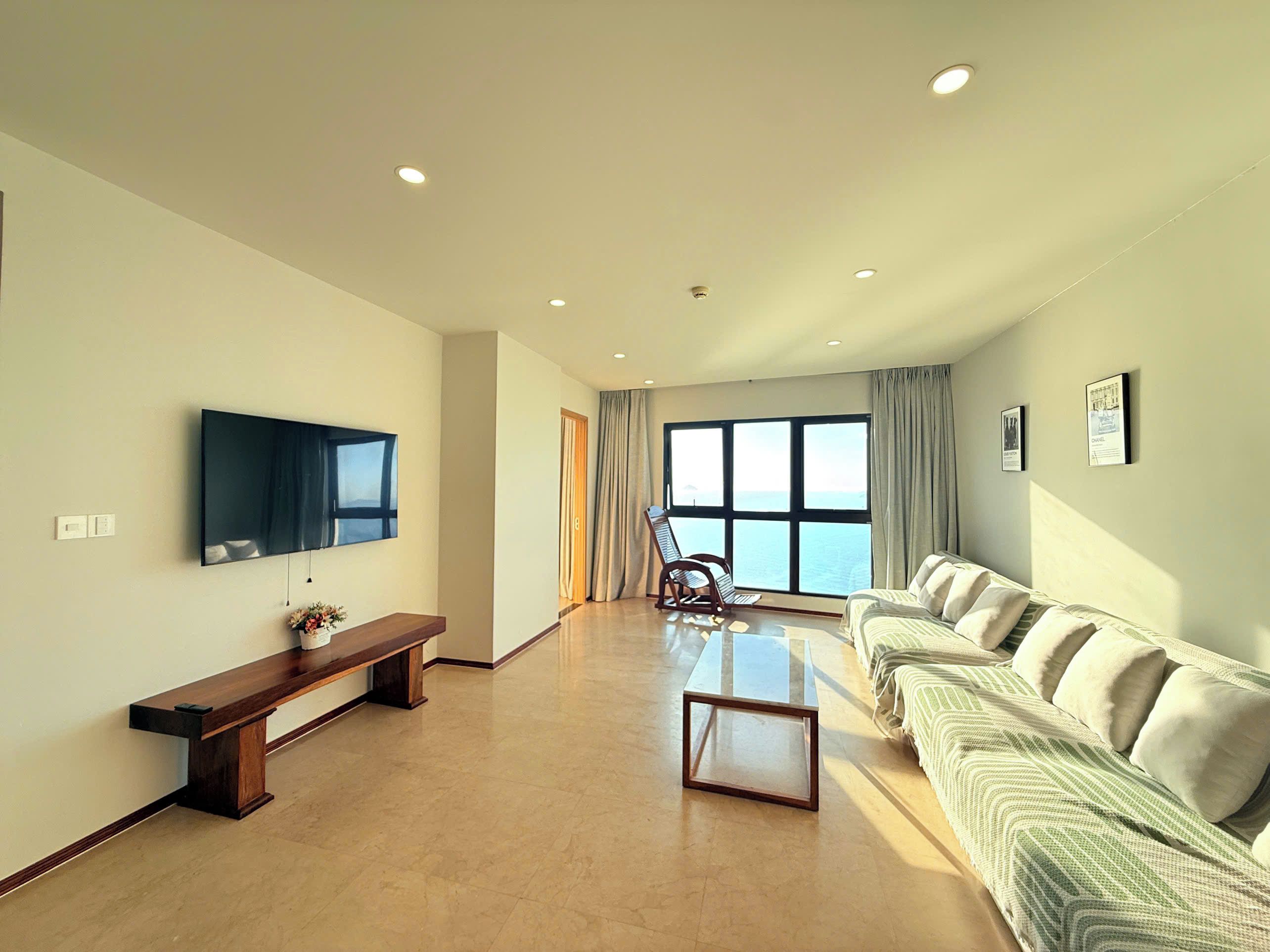 A spacious living room with a large flat-screen TV, a wooden bench, a glass coffee table, and a sofa with cushions.