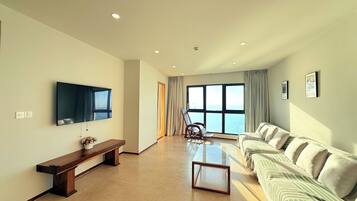 A spacious living room with a large flat-screen TV, a wooden bench, a glass coffee table, and a sofa with cushions.