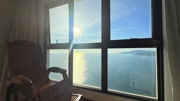 A room with a large window overlooking a body of water and distant land, a wooden chair, and a curtain.