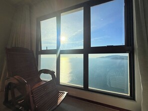 Una habitación con un ventanal grande que da a una masa de agua y tierra a lo lejos, una silla de madera y una cortina.