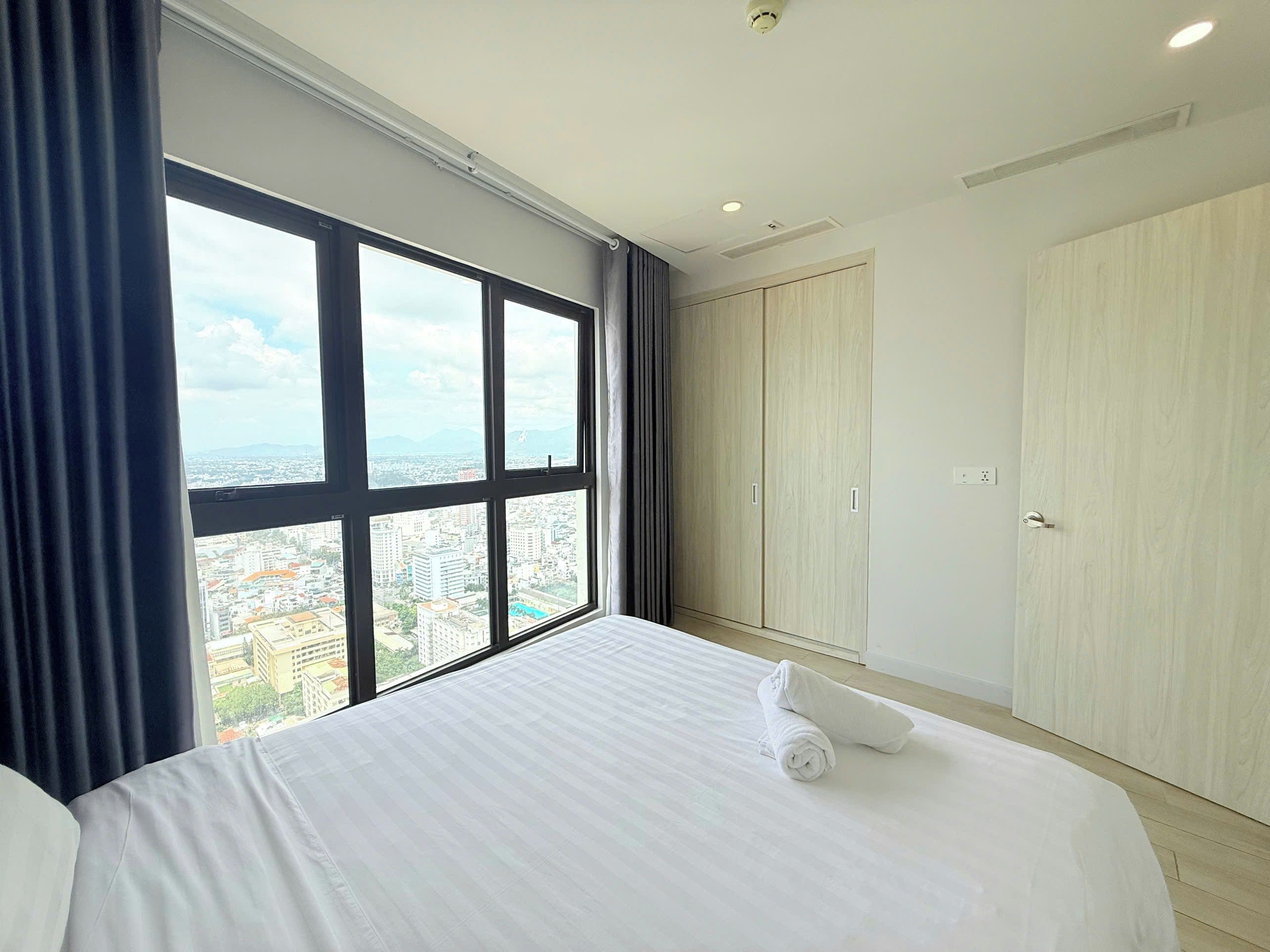 Habitación de hotel con un ventanal grande, cama, armario y vista a la ciudad.