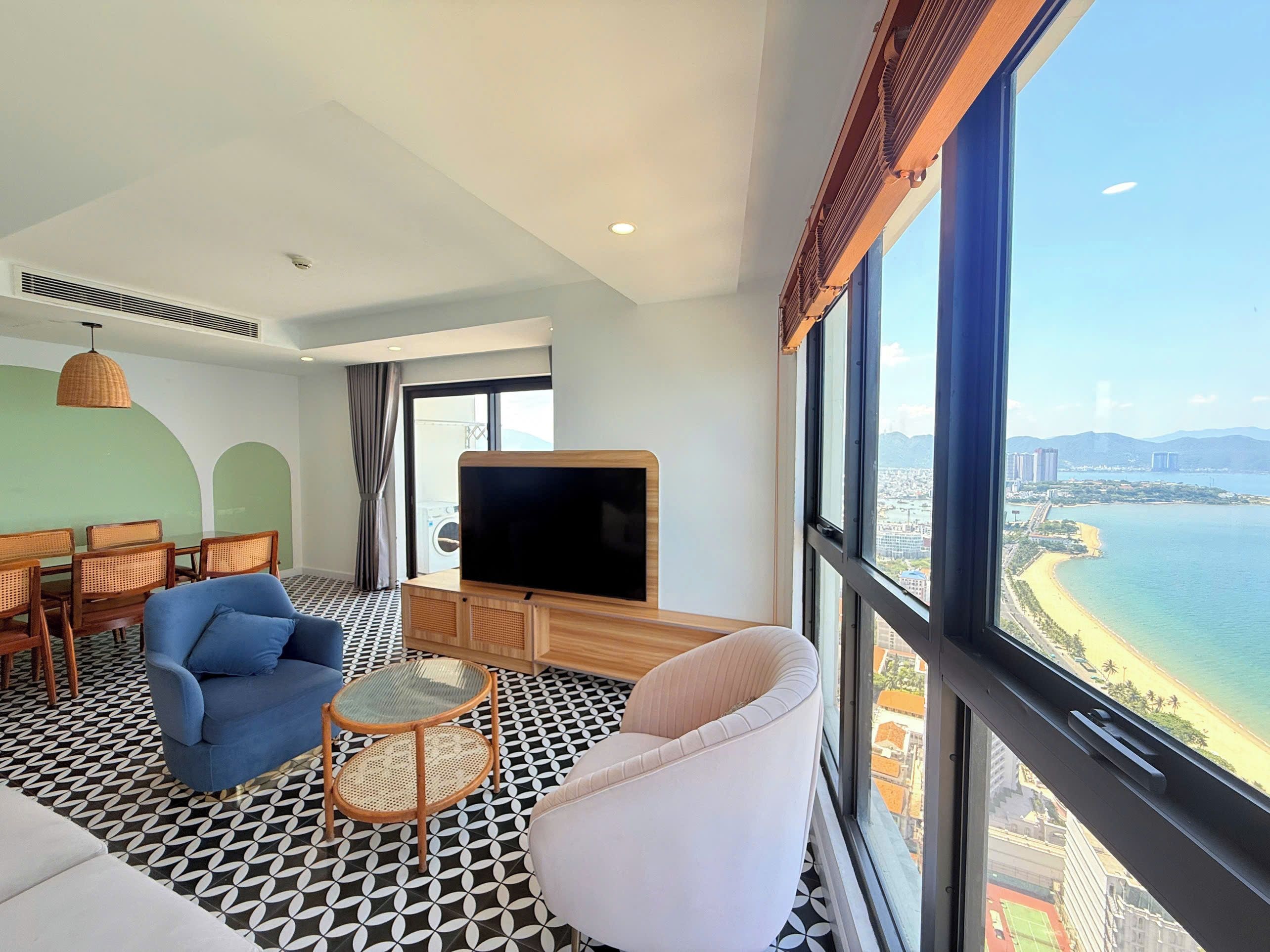 A modern hotel room with a large window, a flat-screen TV, a dining area with a table and chairs, and a patterned floor.