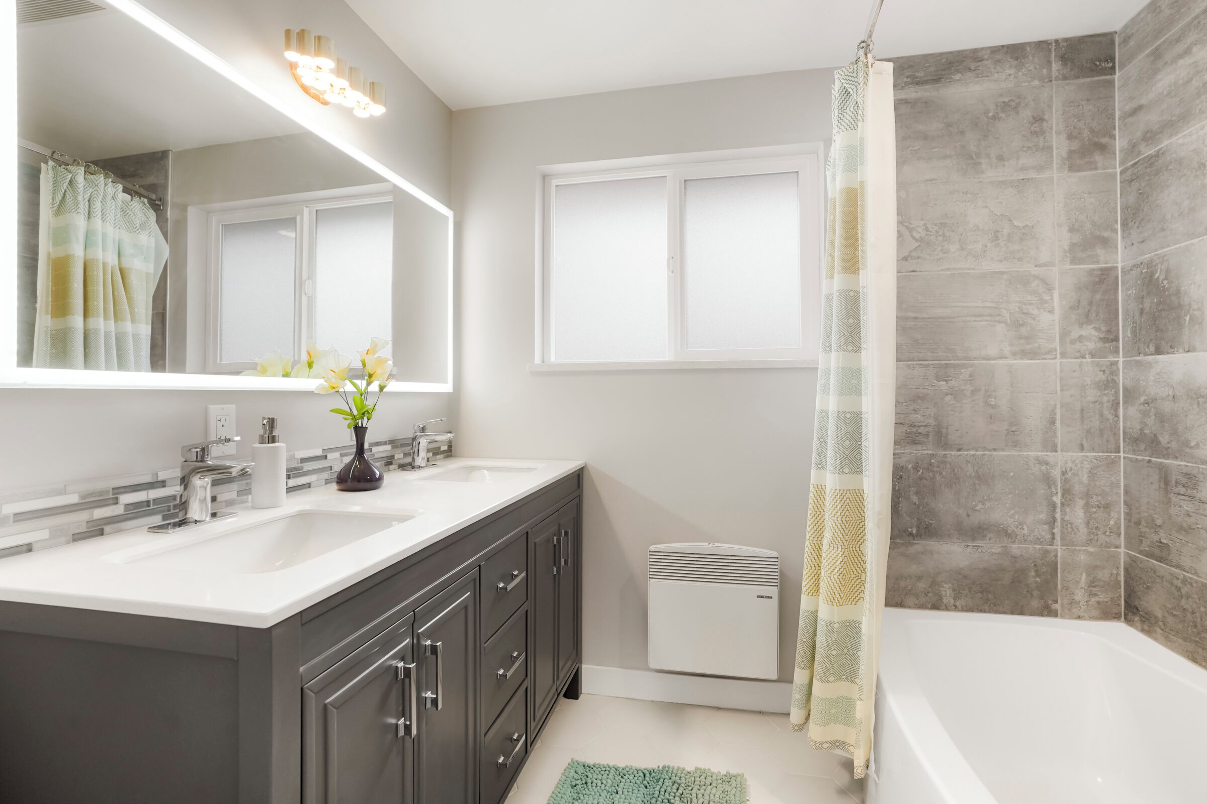 A modern bathroom with a double sink vanity, a large mirror, a shower with a patterned curtain, and a window with frosted glass.