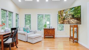 A spacious living room with a large window, a sofa, a wooden coffee table, and a television on a stand. - Dew Drop Inn | 4BR Retreat | Lakeside | Hot Tub (Quinton)