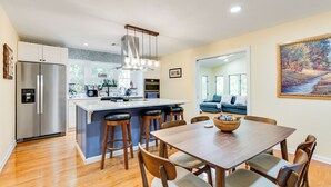 A modern kitchen with a central island, stainless steel refrigerator, and wooden dining table with chairs. - Dew Drop Inn | 4BR Retreat | Lakeside | Hot Tub (Quinton)