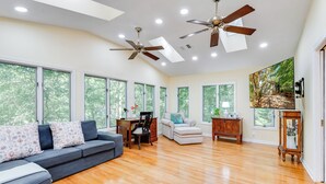 A spacious living room with hardwood floors, a gray sofa, a white armchair, a wooden side table, and a ceiling fan. - Dew Drop Inn | 4BR Retreat | Lakeside | Hot Tub (Quinton)